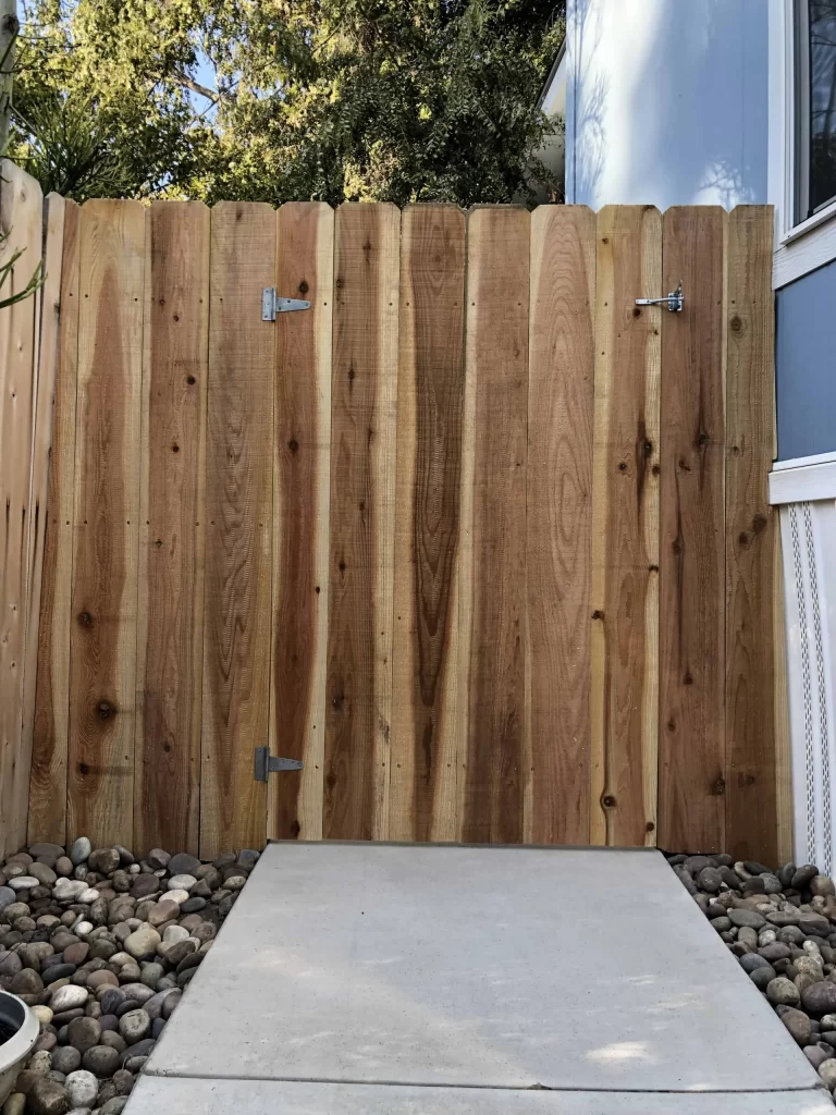 Natural cedar board walk gate with black strap hinges and a latch, opening onto a concrete path between river rock beds
