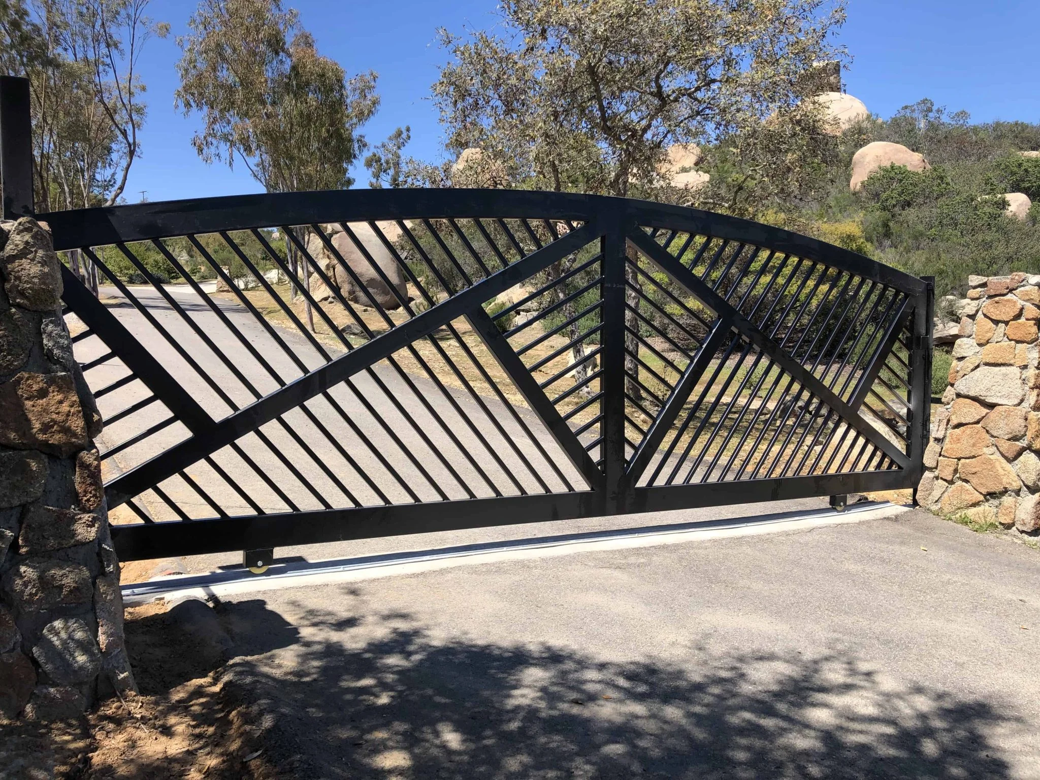 Arched black steel sliding gate with diagonal bars between stone pillars on a hillside driveway in East County San Diego