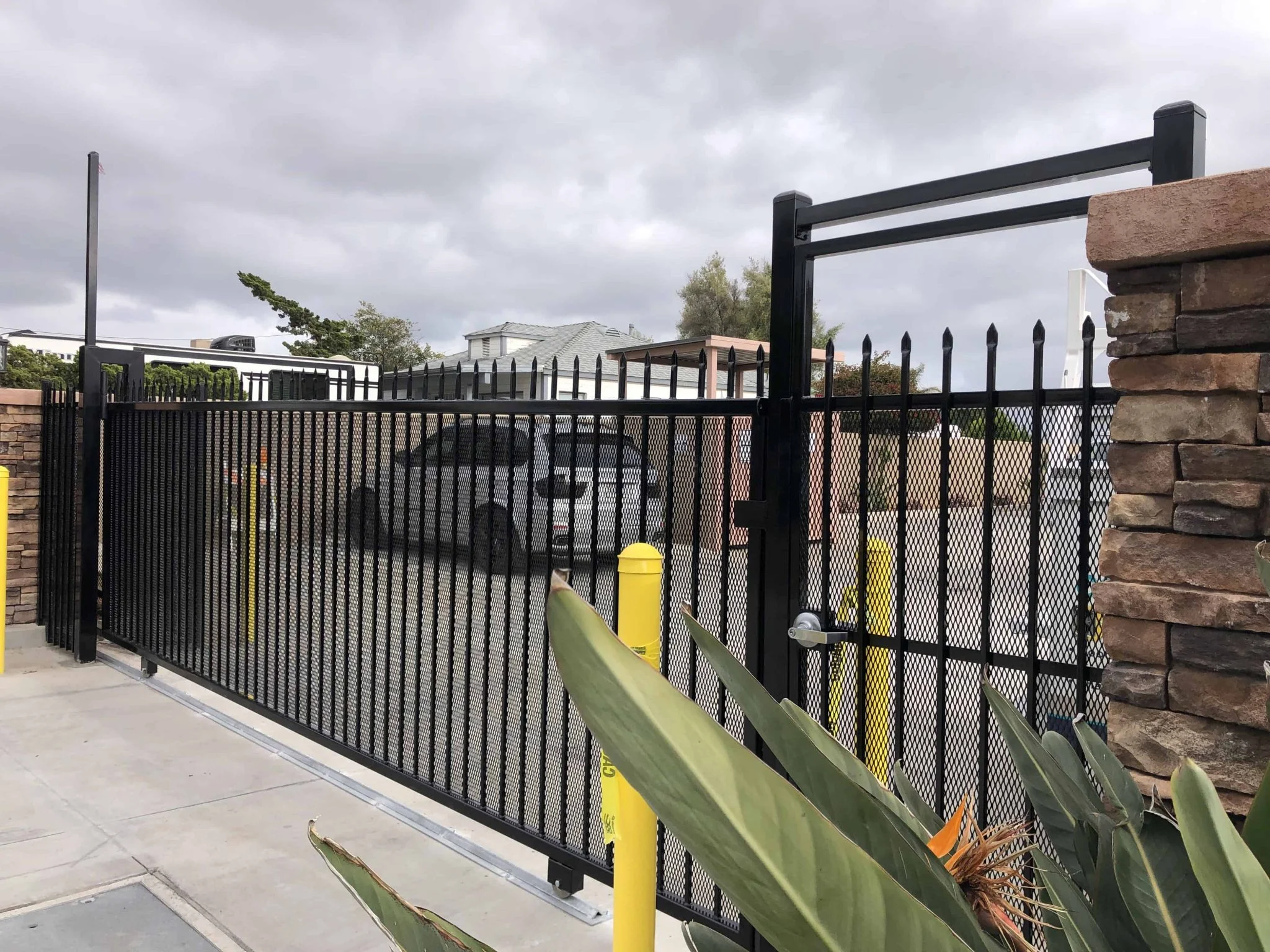Black steel cantilever sliding gate with overhead guide frame beside a stone pillar and yellow bollards