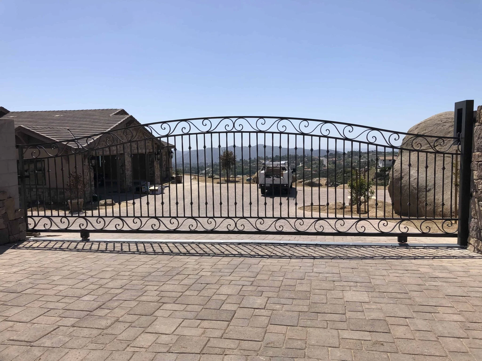 Ornamental wrought iron style sliding gate with scrollwork and an arched top across a paver estate driveway